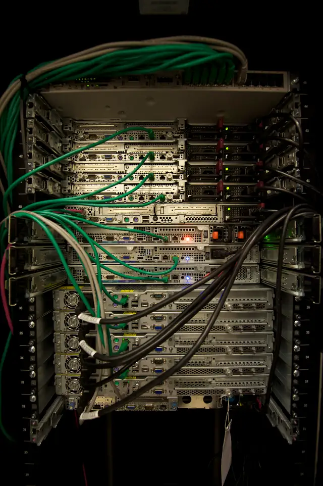 The back of a server rack in a dark room, its servers cabled with green and grey network leads.